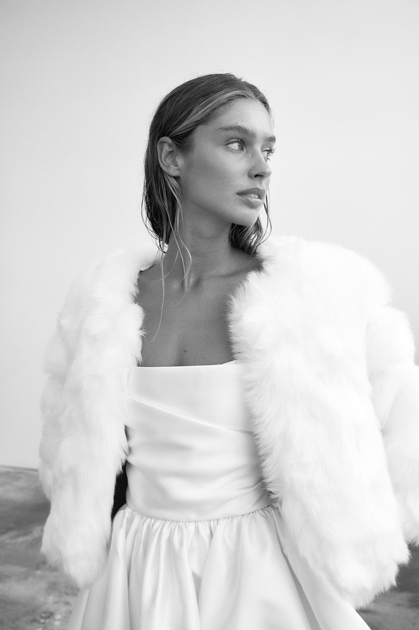 image of bride in black and white looking to the side and wearing a white faux fur jacket with a strapless wedding dress underneath.