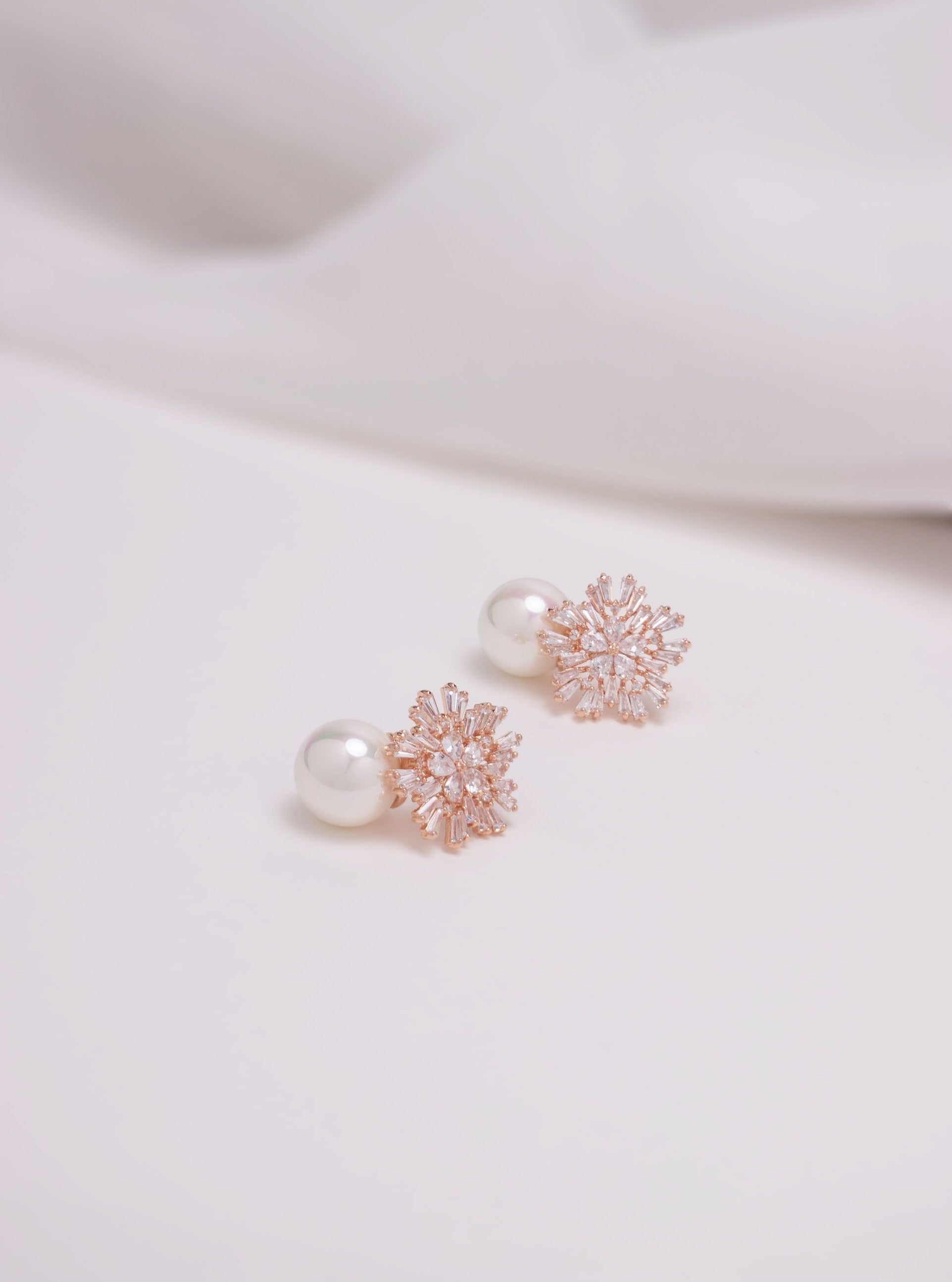Flatlay of rose gold st clair bridal earrings which have A shimmery handset crystal starburst, gently clasps a pearl dropper for a bold and playful yet refined look.