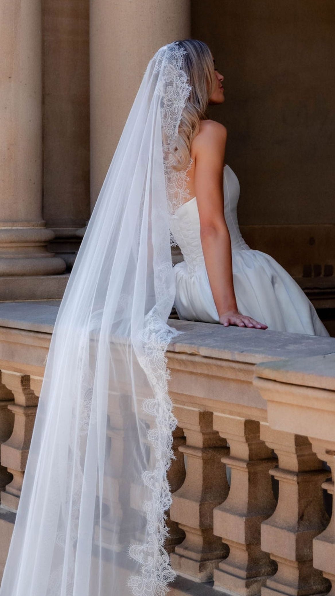 Bride in a white wedding dress with a long veil sitting on a stone bench.