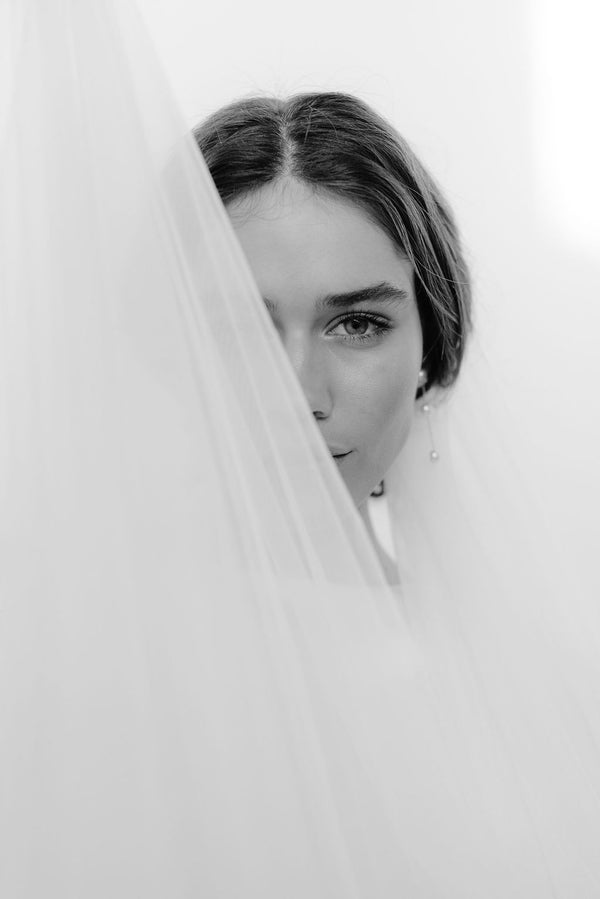 Breathtaking image of a radiant brunette bride looking directly at the camera with a long wedding veil draped diagonally across her face, showcasing the elegance of her bridal ensemble. The delicate fabric drapes gracefully, capturing the timeless beauty of the bride in her short veil wedding.