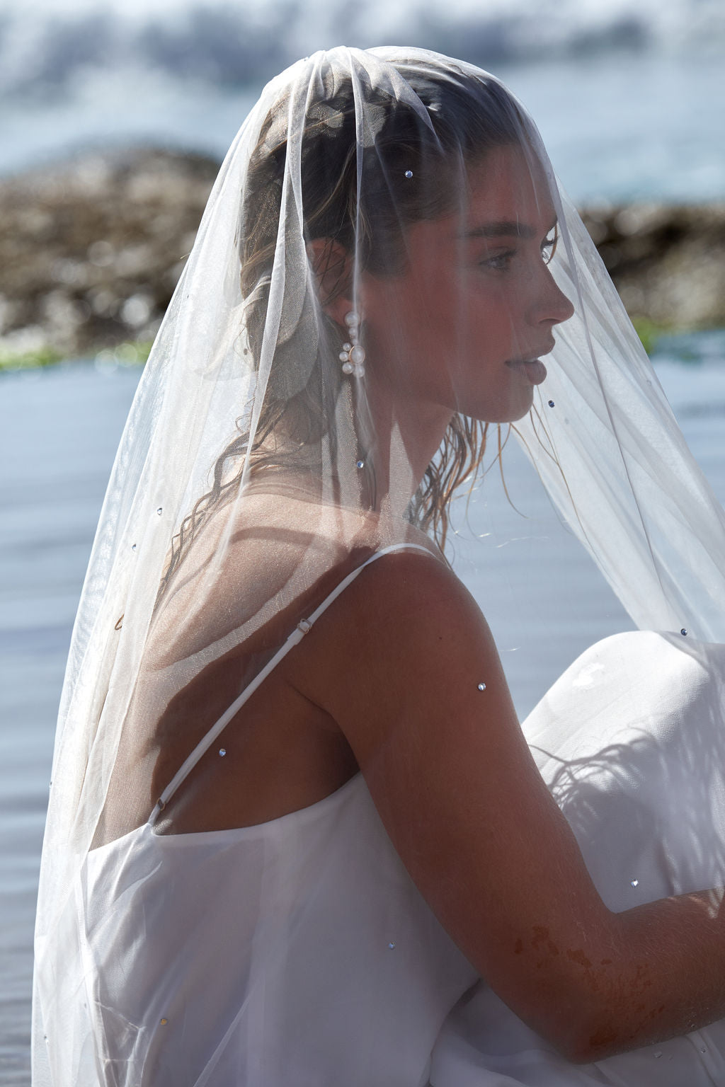 Woman in a wedding dress with a wedding veil sitting by a body of water.