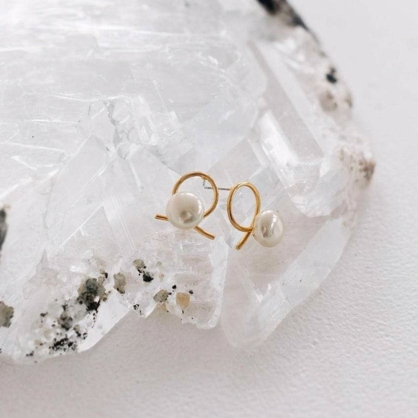 A clear large crystal has a pair of Amelie George Bridal's Airlie earrings on top. They have A beige pearl sits in front of a gold ribbon handtwisted into a knot for modern bridal looks