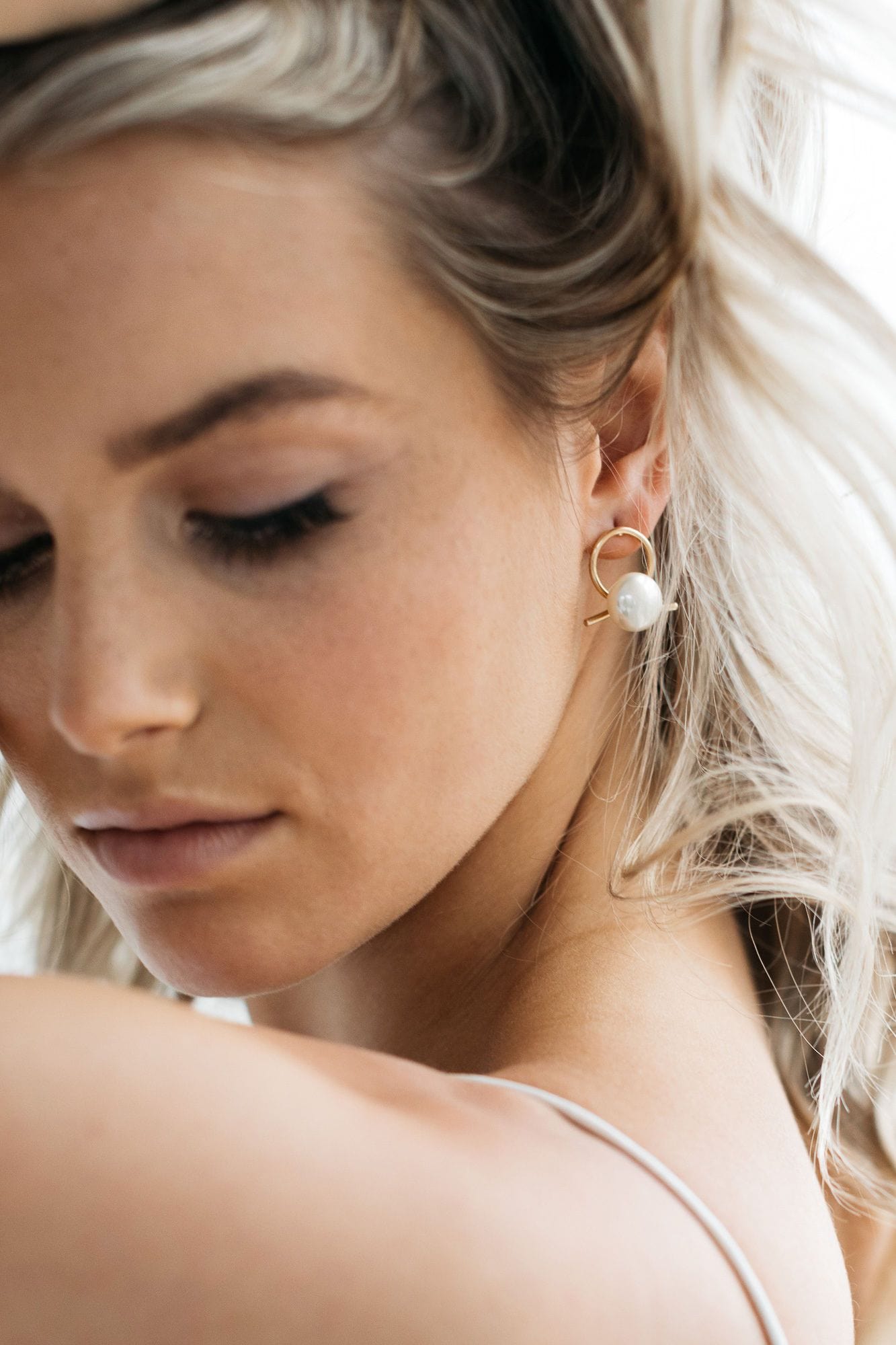 A model wears Airlie earrings which have A beige pearl sits in front of a gold ribbon handtwisted into a knot for modern bridal looks. The model is lifting her blonde hair