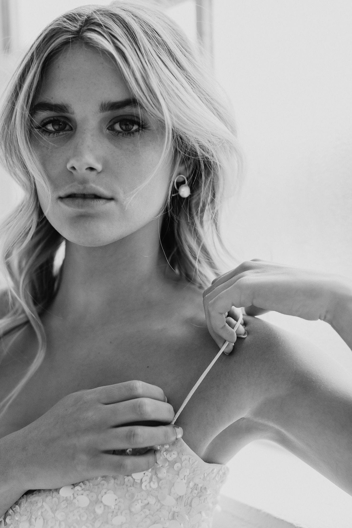 Black and white image of bride with natural wavy blonde hair for wedding photography wearing Airlie earrings which have A beige pearl sits in front of a gold ribbon handtwisted into a knot for modern bridal looks